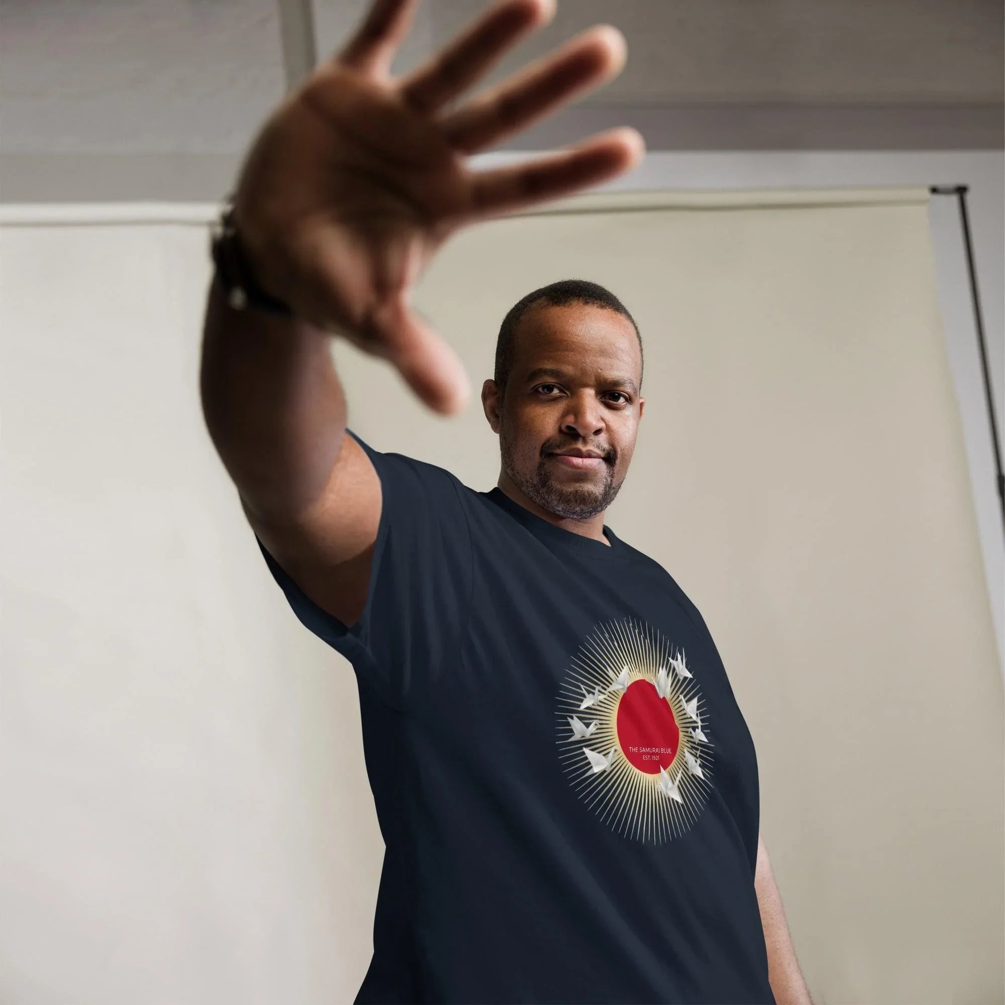 Man wearing navy t-shirt with red sun and white cranes design, hand raised in greeting