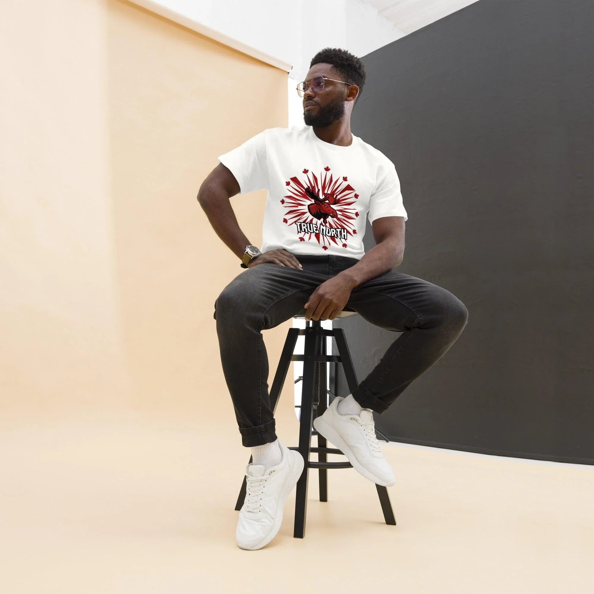 Man wearing white True North moose graphic tee, black jeans, white sneakers, sitting on stool